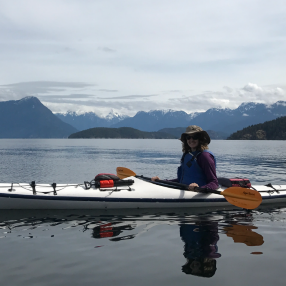 Kaitlyn smiling at the camera while sitting in a kayak that is in a large body of water. Mountains are in the distant background.